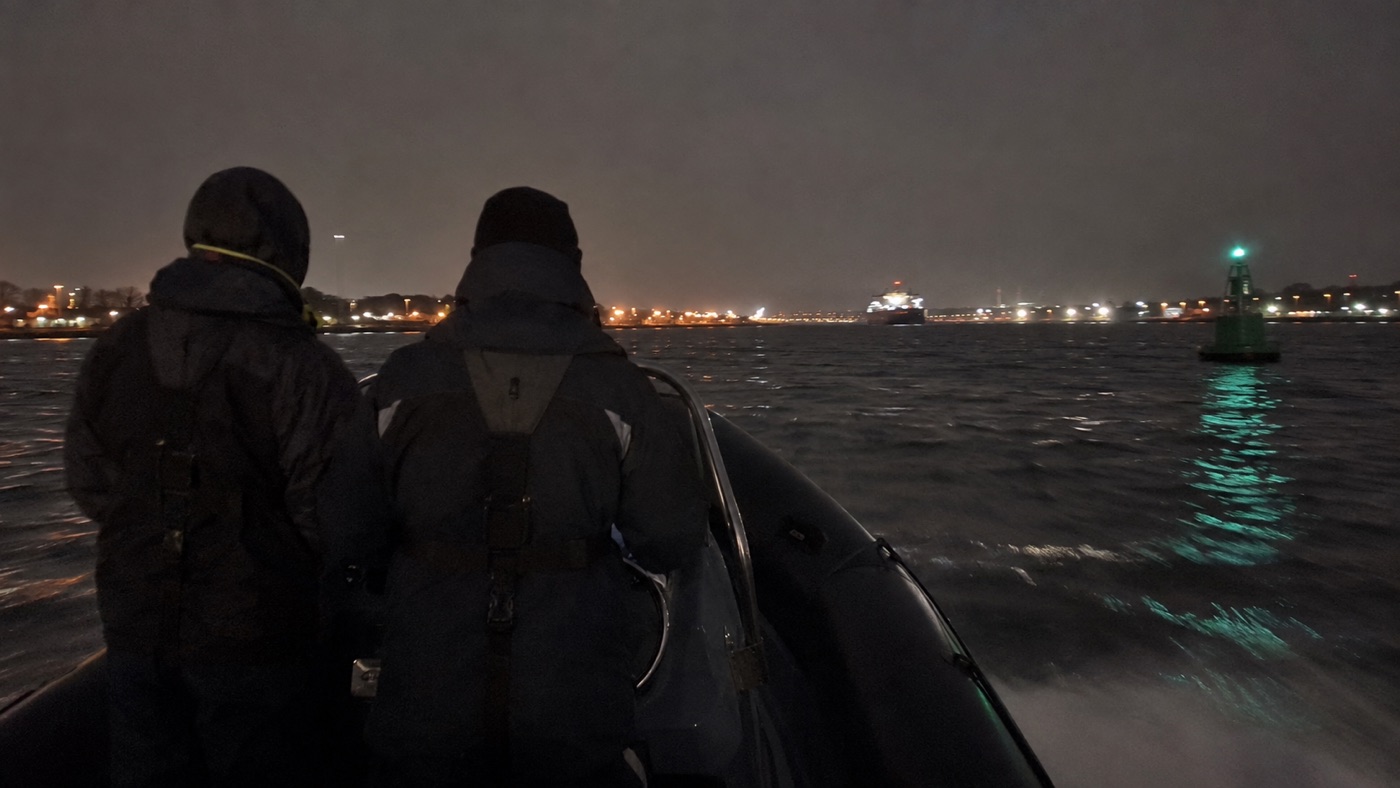 Two crew running a RIB at night towards a lit shoreline with a green channel buoy to starboard
