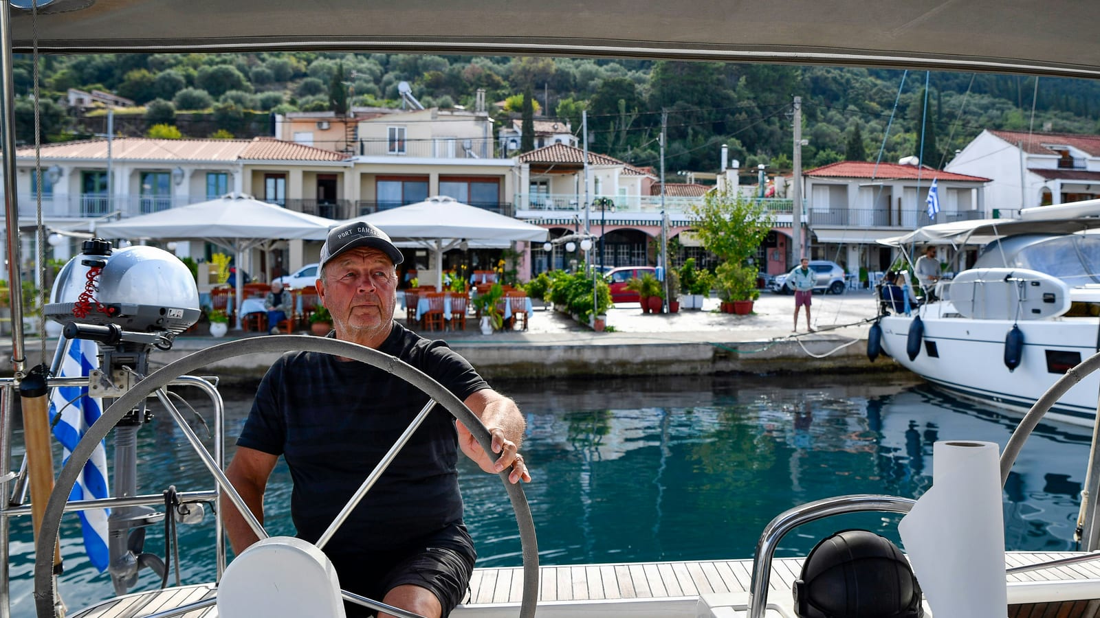 Skipper at the wheel of a yacht stern-to on a harbour quay with a waterfront village behind
