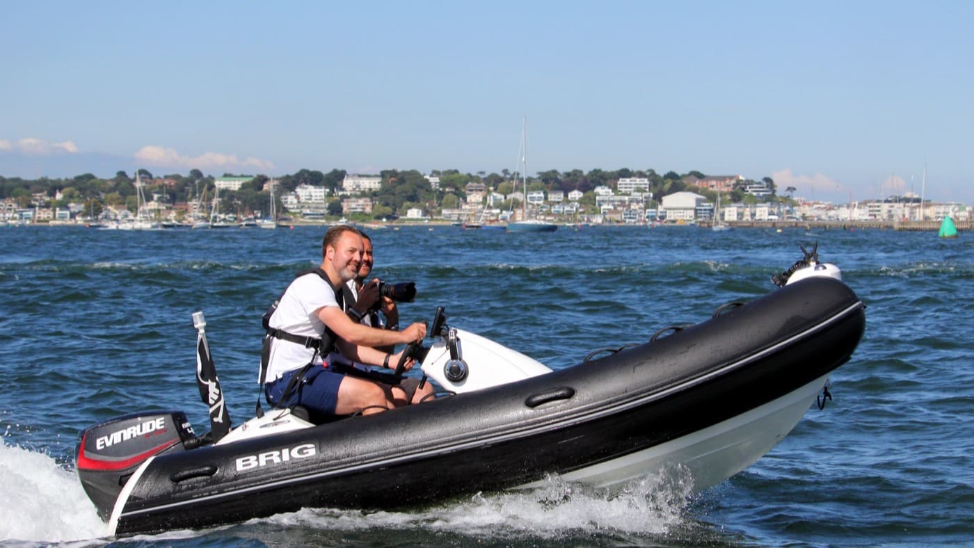 Two men in lifejackets driving a small RIB at speed across a sheltered harbour on a sunny day