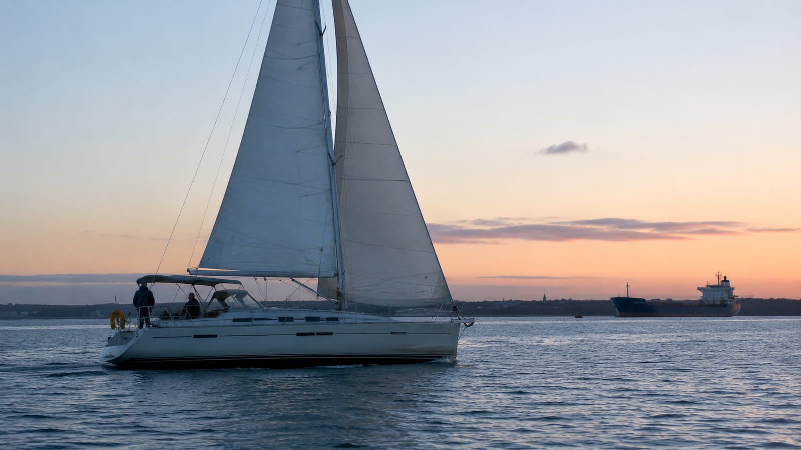 Cruising yacht under sail at dusk with a cargo ship on the horizon